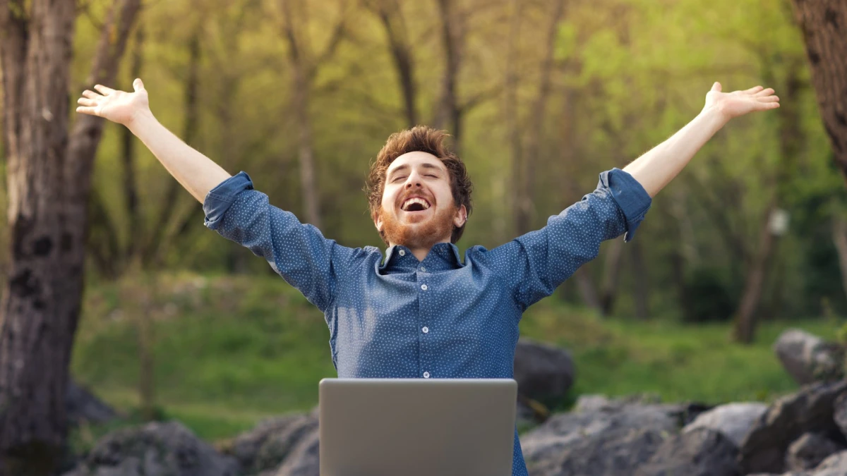 A man with a laptop raising his arms in a green forest