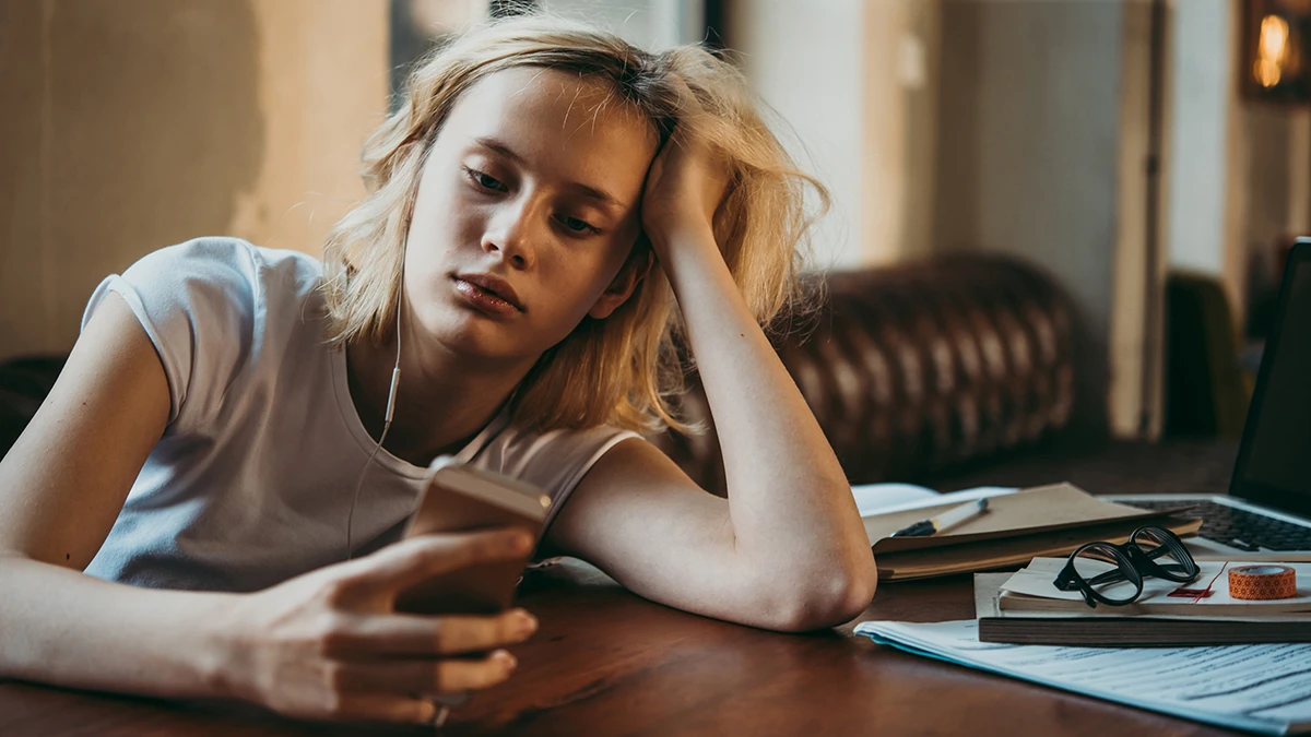 A woman holding a phone and resting her head on her hand at a desk with papers and a laptop