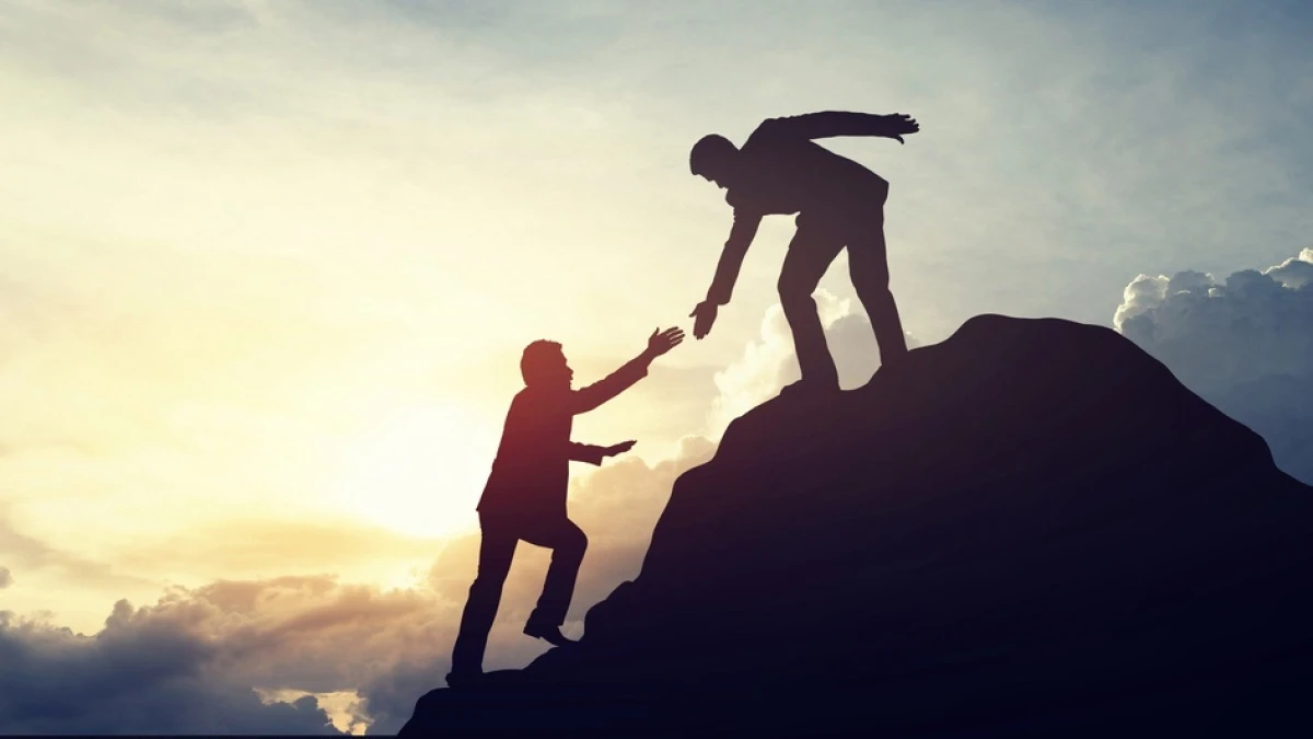 Silhouettes of two people on a rock reaching towards each other against a bright sky