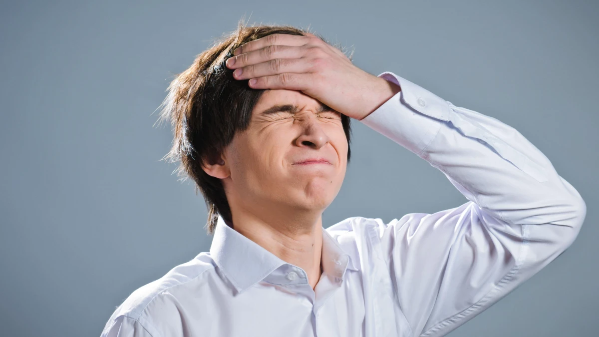 A man in a white shirt with closed eyes and a hand on his forehead