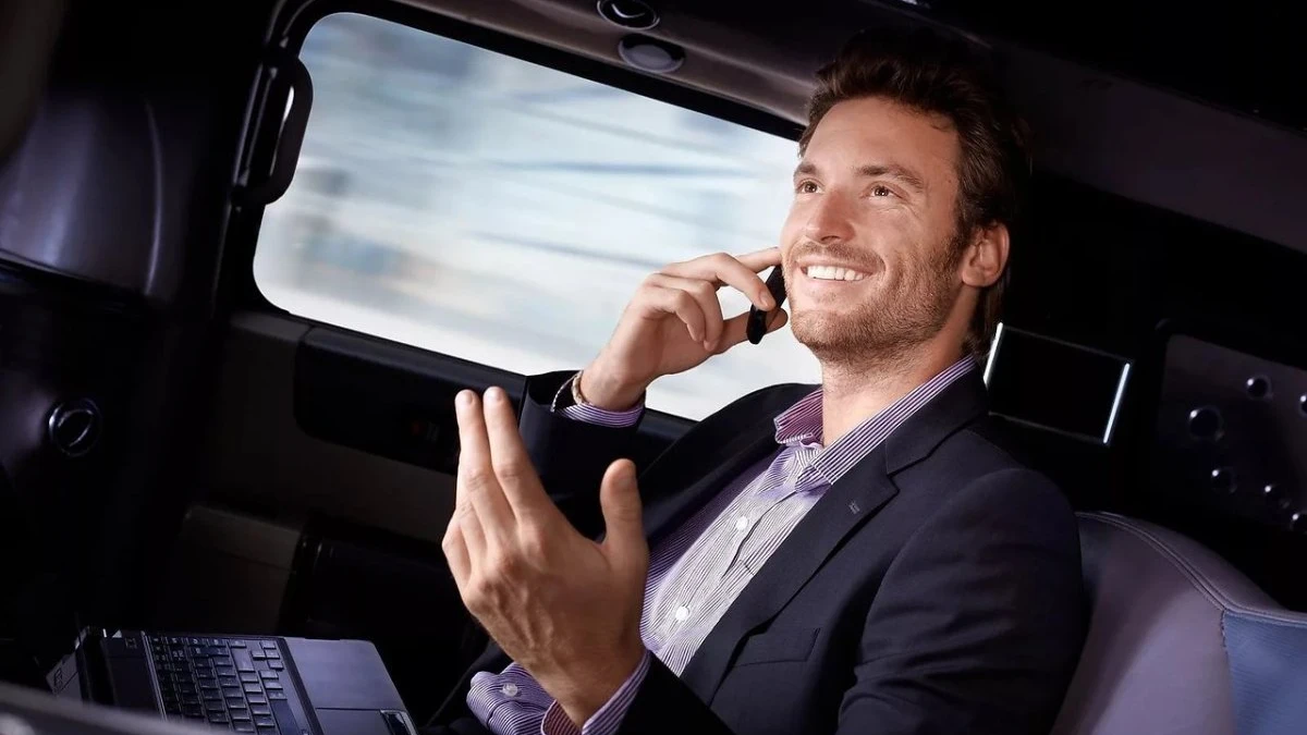 A man talks on a phone in a car with a laptop on his lap