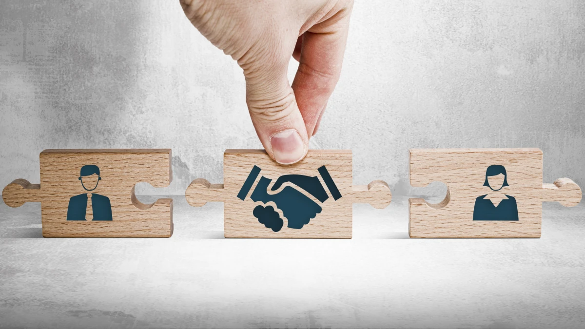 Wooden puzzle pieces showing two people and a handshake, with a hand holding the centre piece