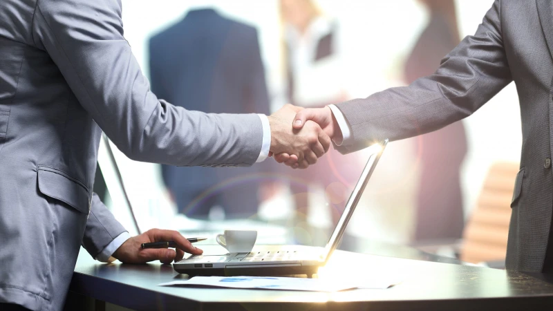 Two people in business suits shaking hands across a table with a laptop