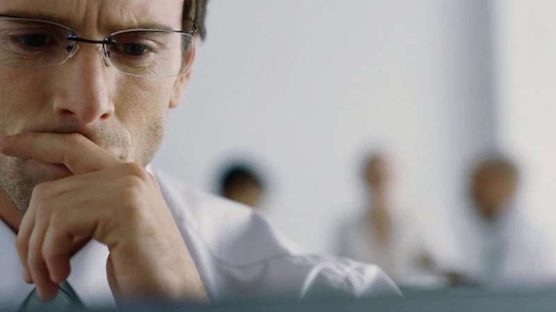 A man wearing glasses holds a hand to his chin with people blurred behind him in an office
